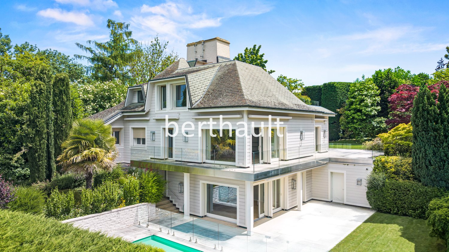 Two-story house with a white exterior, a tiled roof, and large windows. The house is surrounded by lush greenery, including palm trees and other vegetation. There is a swimming pool in the backyard and a balcony or terrace on the upper level.