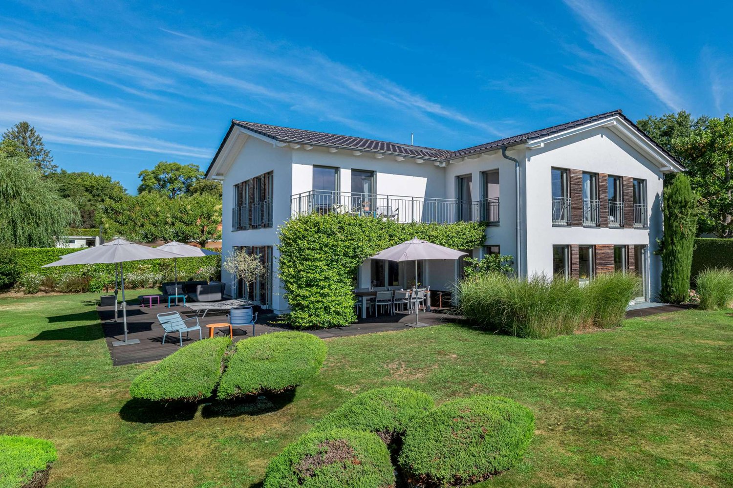 Two-story white house with balconies, surrounded by a well-maintained garden with trees and shrubs. There is a patio area with outdoor furniture in the backyard. The house has a tiled roof and large windows.