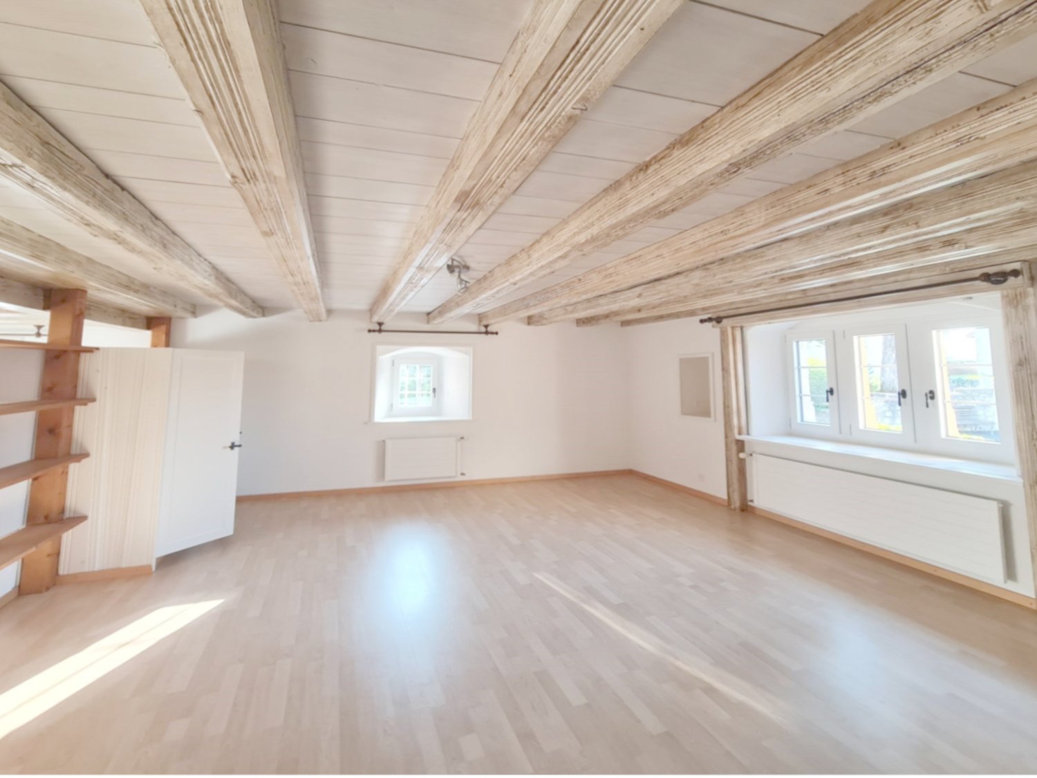 Large open room with wooden beamed ceiling, white walls, hardwood flooring, and built-in shelving units. The room has several windows providing natural light.