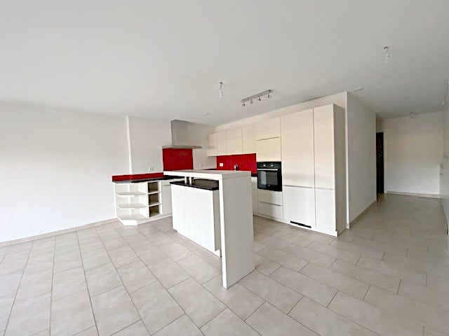 Spacious open-plan kitchen with white cabinets, red accents, and modern appliances including a dishwasher. The kitchen has a tiled floor and appears to be part of a larger living space.