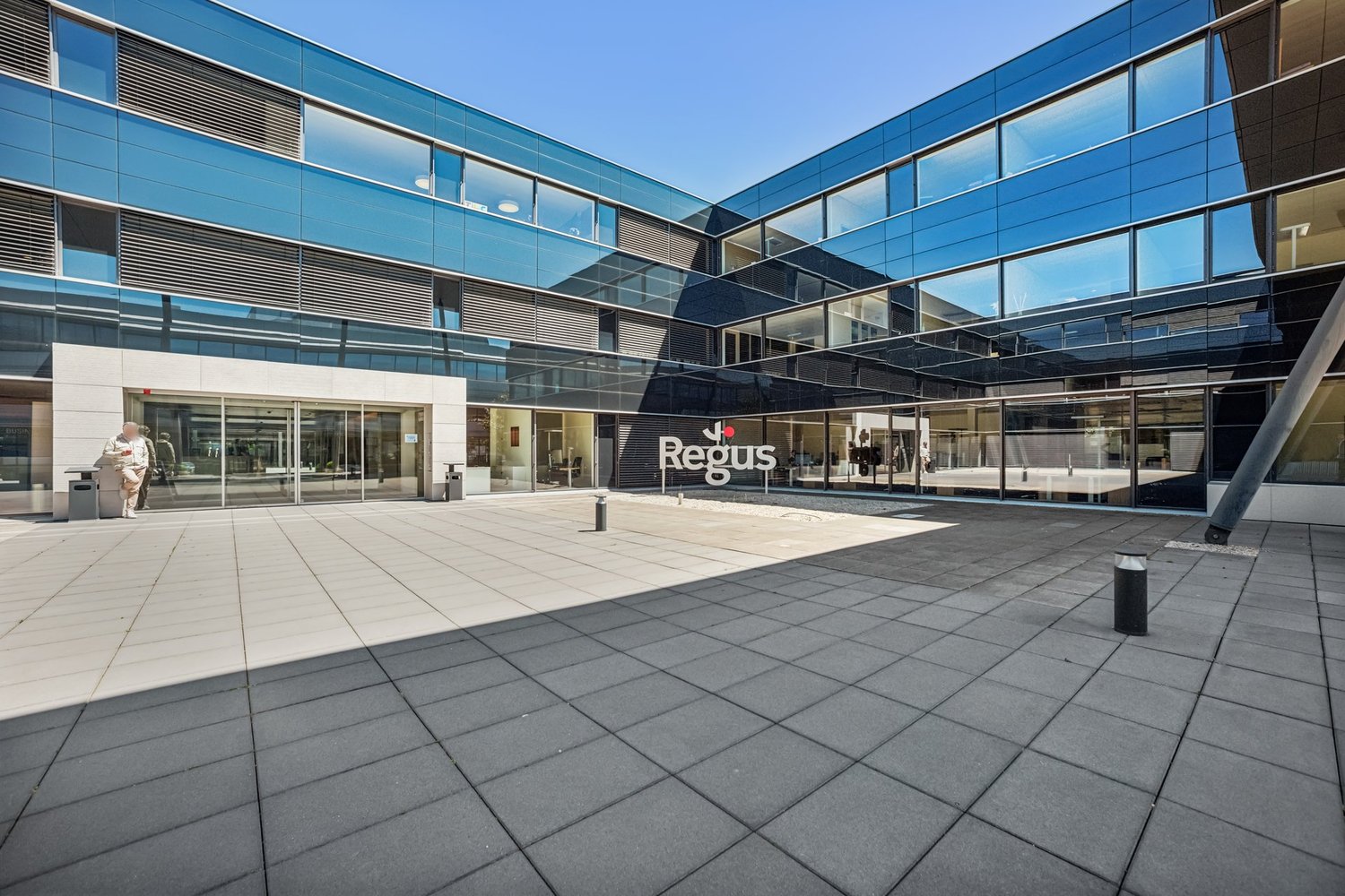 Modern glass and steel commercial building with the Regus logo prominently displayed. The building has a large open plaza area with paved flooring in front of the main entrance.
