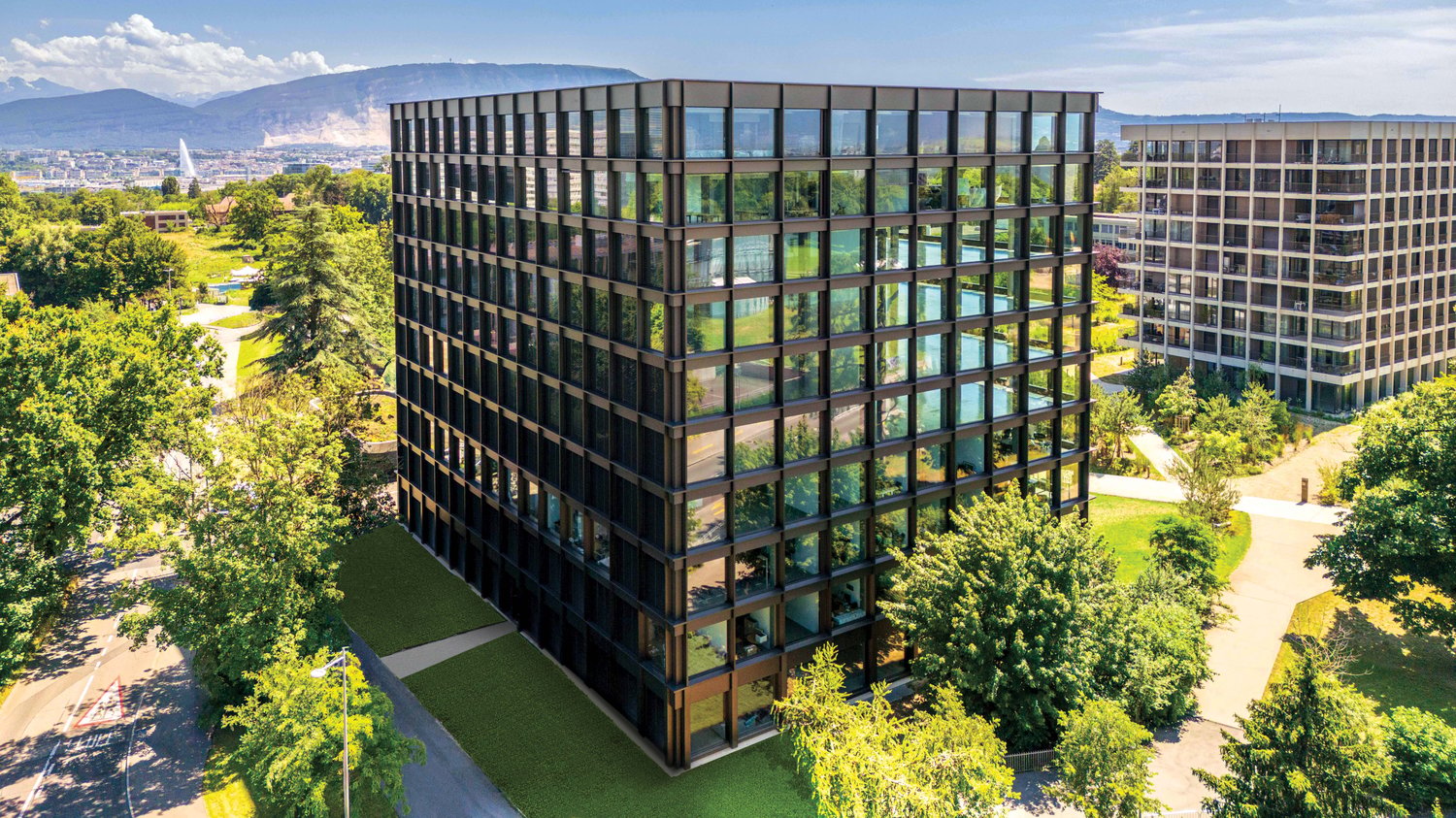 A modern, multi-story office building with a glass facade surrounded by lush greenery and landscaping. The building has a rectangular shape with numerous windows and appears to be located in an urban setting with mountains visible in the background.