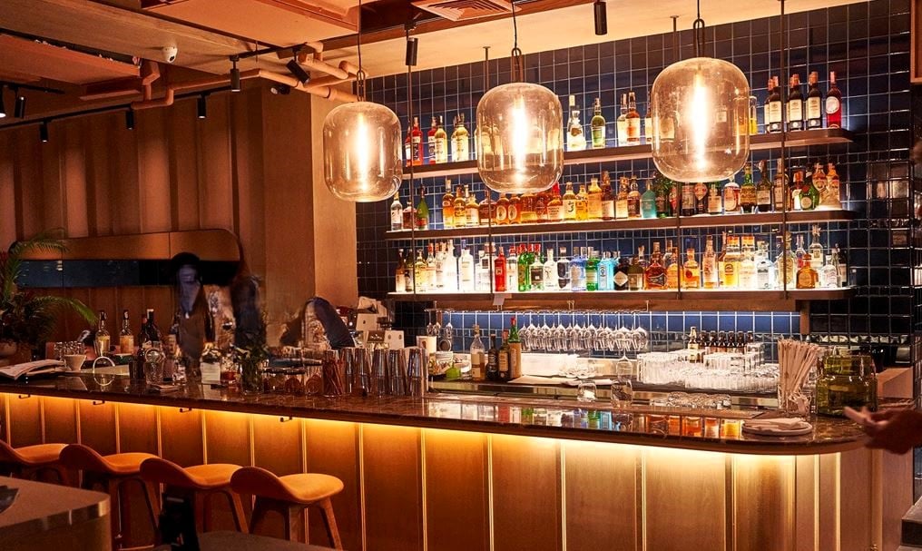 Well-stocked bar with a variety of liquor bottles, glassware, and large pendant lights hanging above the bar counter. The bar has a modern, cozy atmosphere with wooden accents and tiled walls.