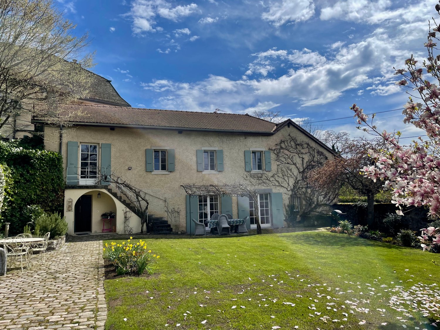 Two-story stone house with green shutters, surrounded by a well-maintained garden with a cobblestone driveway, flowering trees, and outdoor seating area