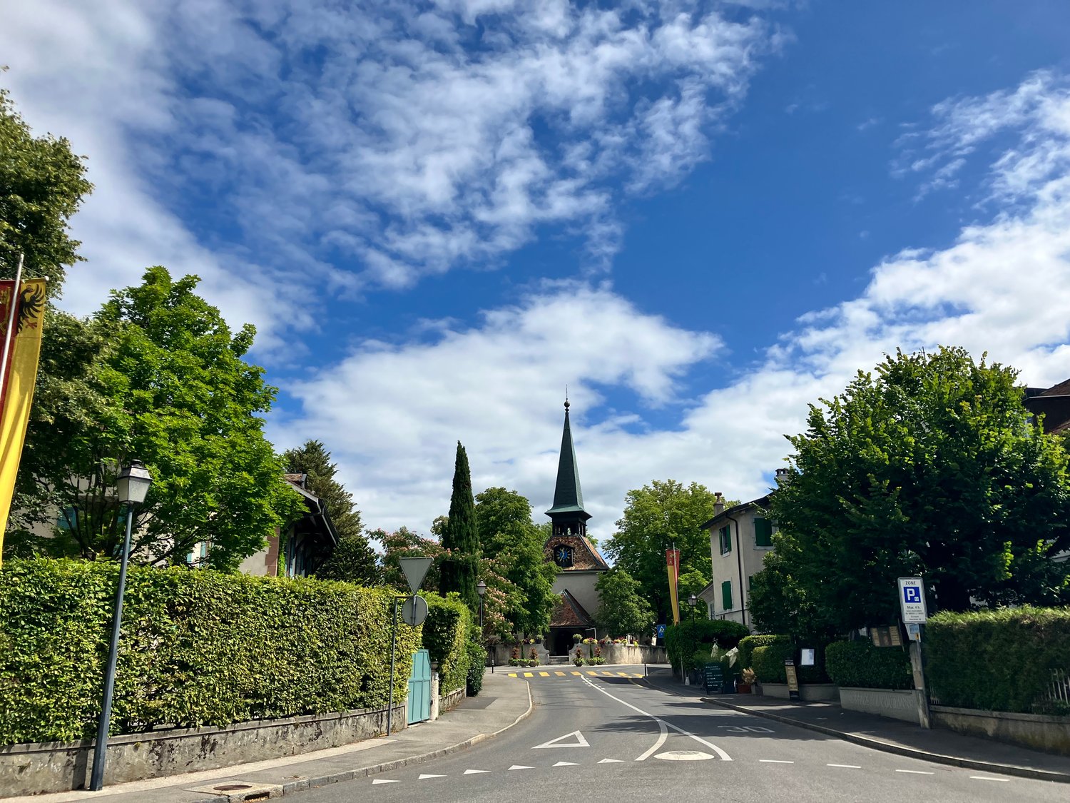 The image shows a picturesque street in a small town or village. The scene features a church steeple with a green roof, surrounded by lush green trees and hedges. The street is lined with buildings and has a parking area visible. The sky is blue with fluf