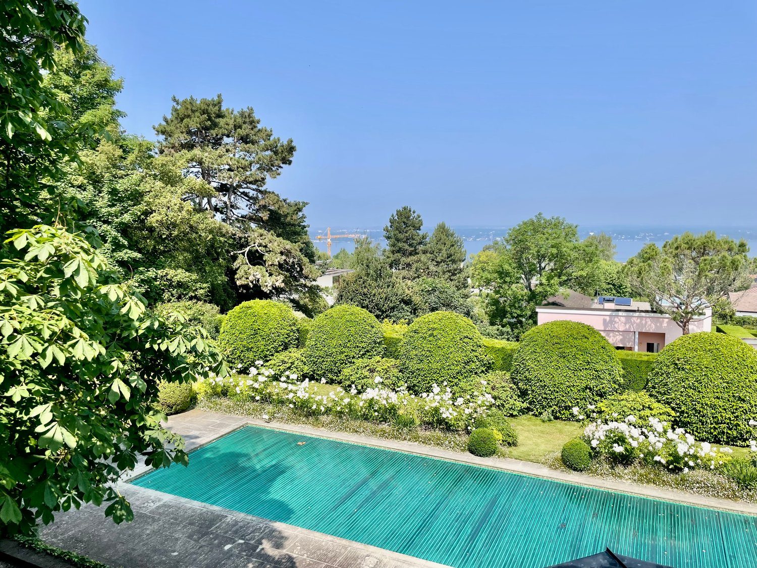 Lush, well-manicured garden with a swimming pool, surrounded by green trees and shrubs. The garden features neatly trimmed hedges and a clear blue sky in the background.