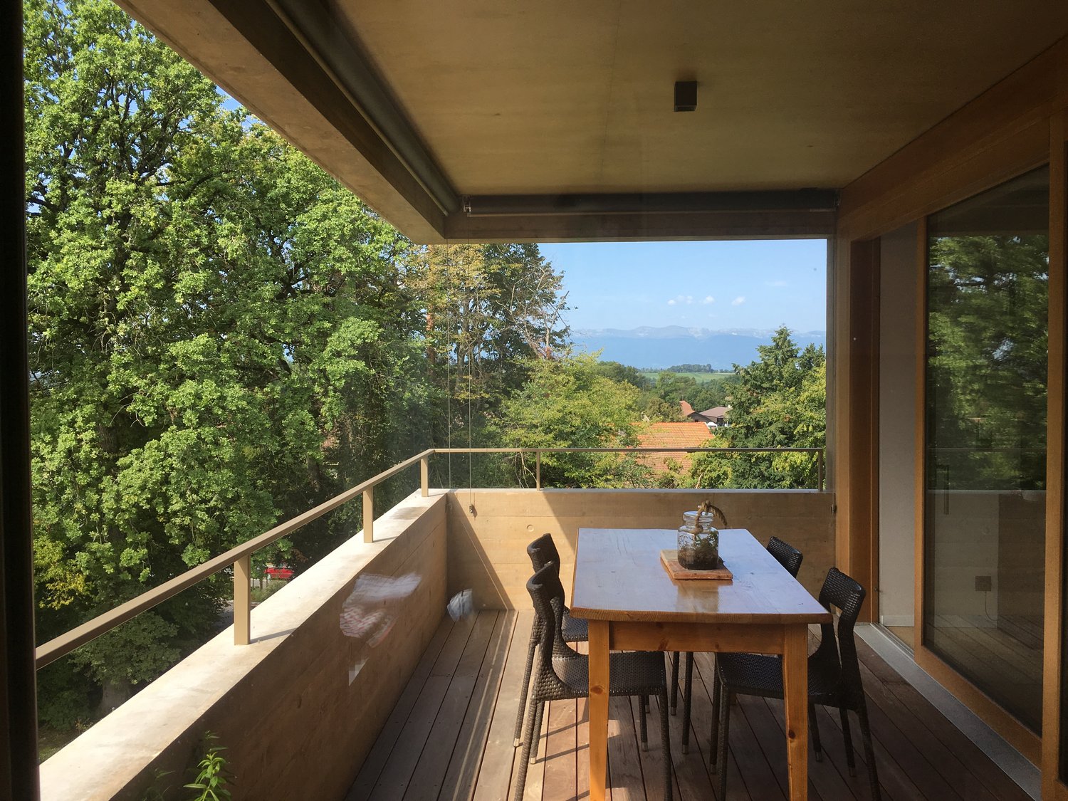 Covered balcony with wooden floor, glass railing, and a dining table with chairs overlooking a scenic view of trees and distant buildings