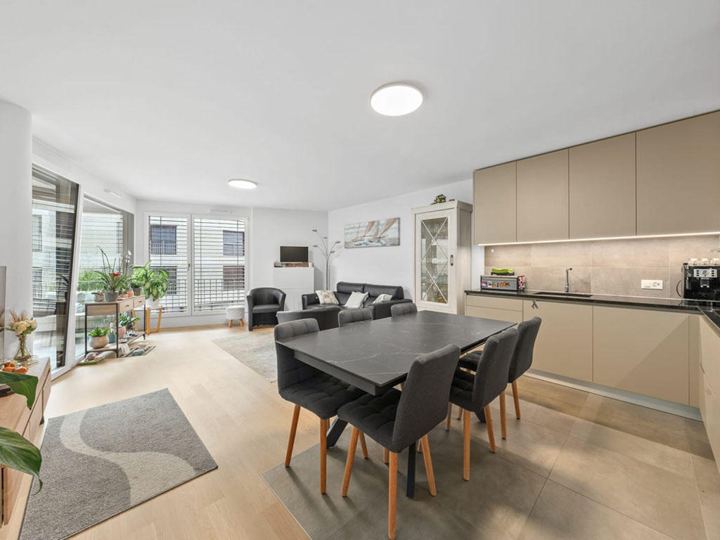 Modern living room with dining area, wooden floor, large windows, modern furniture, and kitchen area.