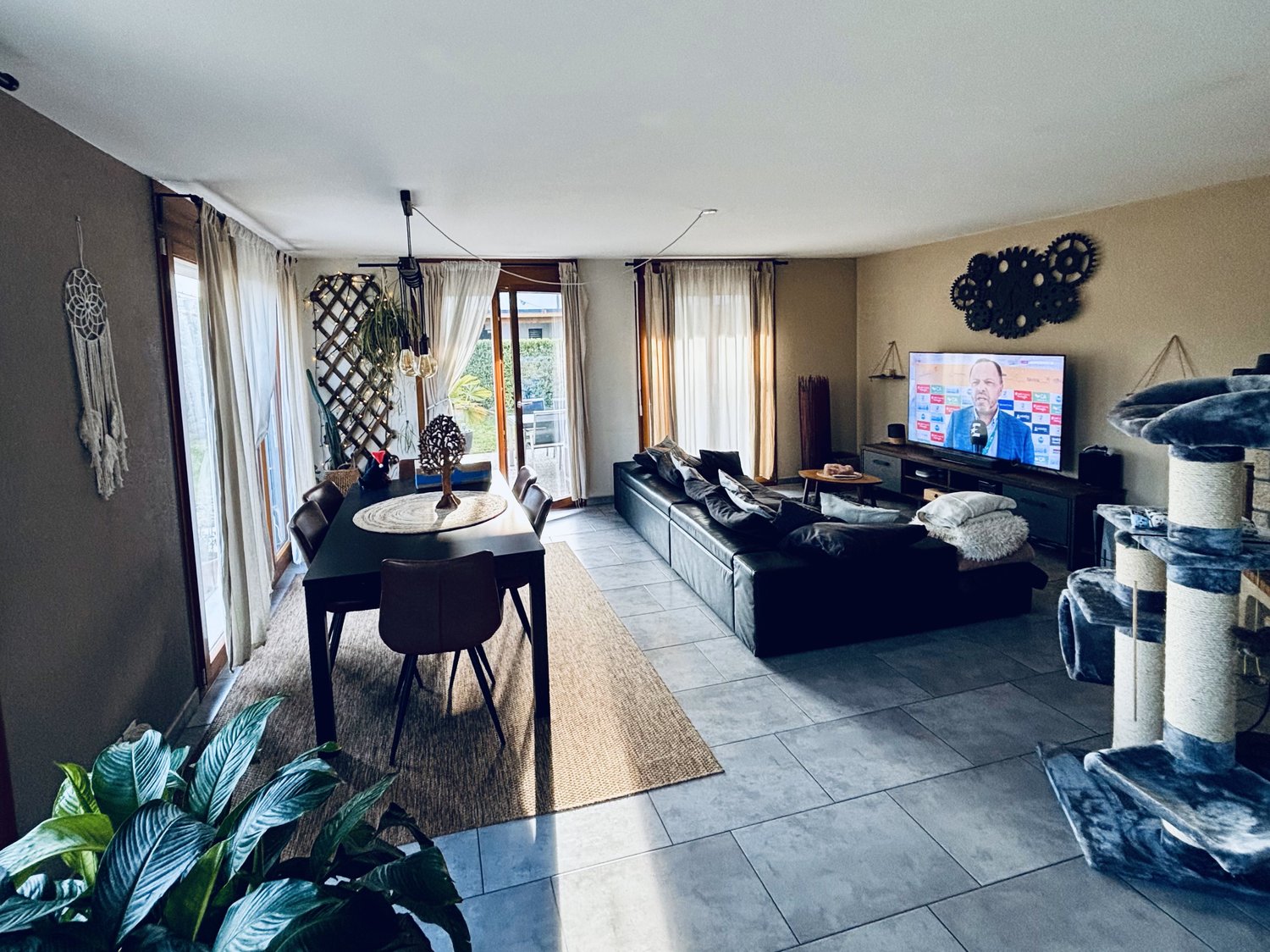 Open-concept living room and dining area, with large windows, black leather sofa, TV stand, dining table and chairs, decorative wall art, and potted plants