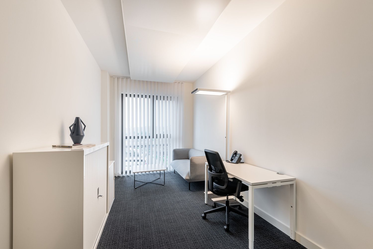 Bright and modern office space with large windows, white walls, and gray carpeting. The room features a desk with a computer, a sofa, and a storage cabinet. There is also a vertical blind covering the window, providing privacy and light control.