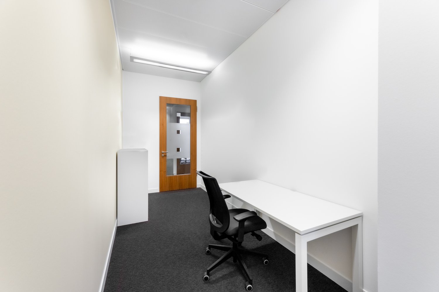 The image shows a small office space with a white desk, a black office chair, and a wooden door with a window. The room has white walls and a gray carpeted floor. The lighting appears to be provided by overhead fixtures.