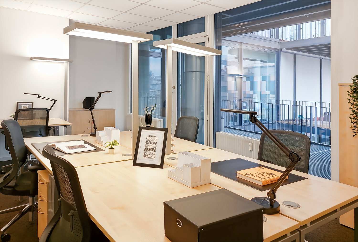 Modern and well-lit office space with large windows providing a view, desks with computers and office equipment, framed artwork on the walls, and various decorative elements on the desks.