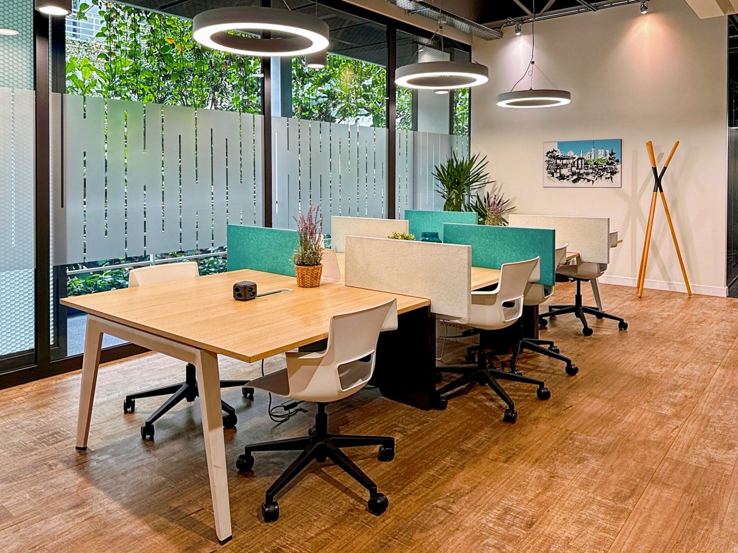 Open office space with wooden tables, office chairs, hanging pendant lights, plants, and a large window overlooking a balcony or terrace area