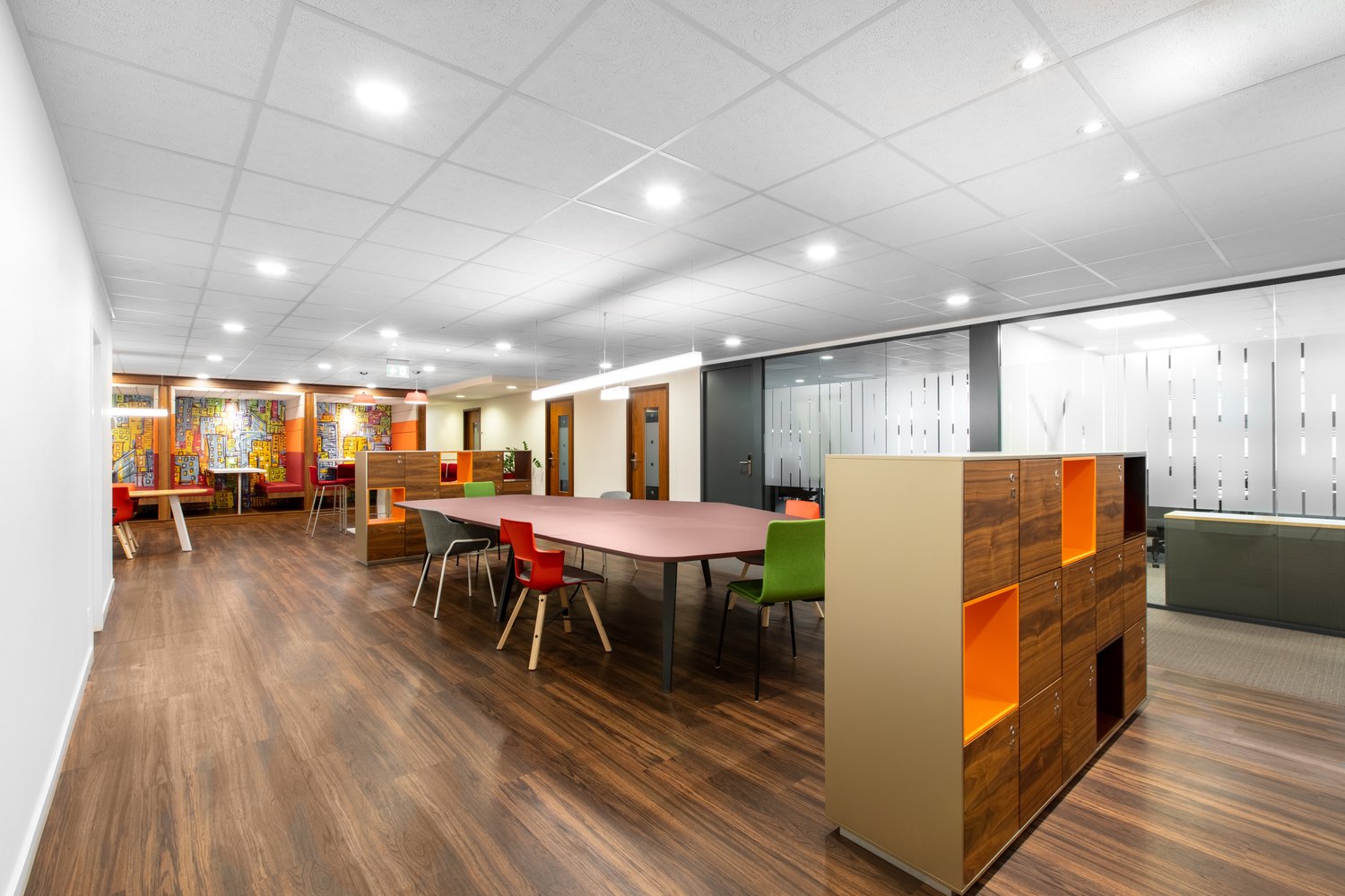 Large open office space with hardwood flooring, white ceiling tiles, and colorful furniture including a long conference table, chairs, and storage units. The space appears to be a modern and vibrant office environment.