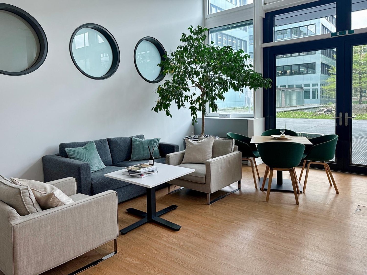 A spacious living room with wooden floors and white walls. The room includes a sofa with pillows, a coffee table, a table with four chairs, a tree potted plant, and windows with a view outside to a building and lawn.