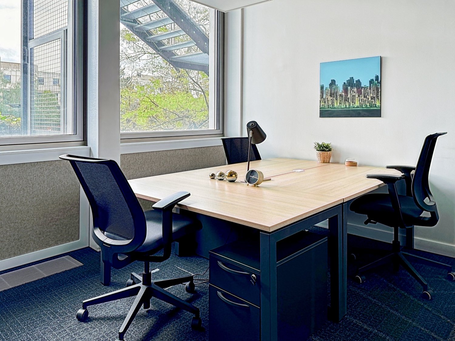 Modern office space with large windows overlooking greenery, wooden desk, black office chairs, and a cityscape painting on the wall.
