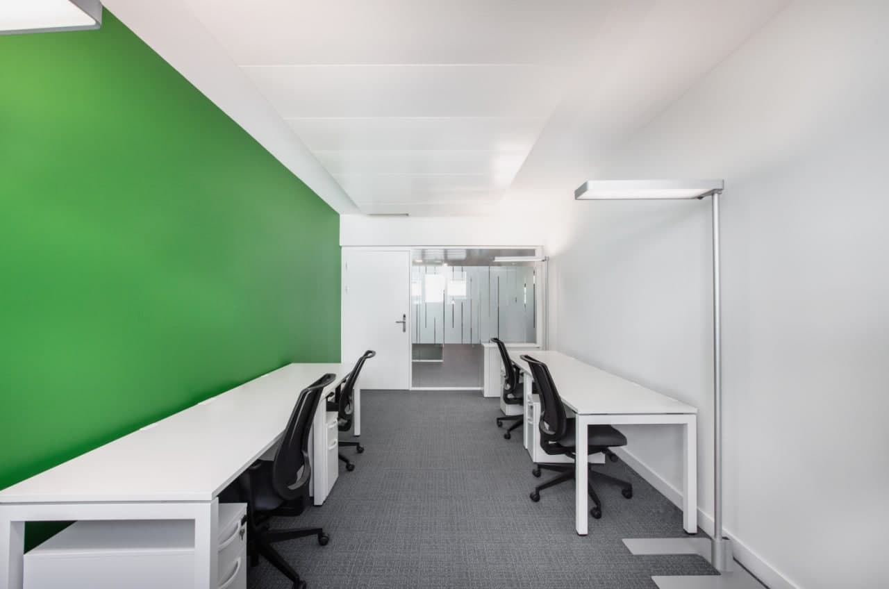 The image shows a modern office space with a green accent wall, white desks, black office chairs, and a carpeted floor. The space appears to be well-lit with overhead lighting fixtures. There is a glass-paneled door visible in the background, leading to a