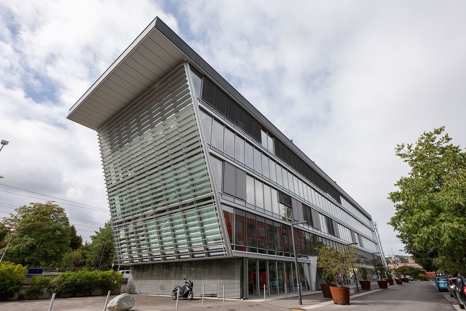 A modern, multi-story commercial or office building with a distinctive angular glass and metal facade. The building has large windows and a sleek, contemporary design. There are trees and other greenery surrounding the building, suggesting it is located i