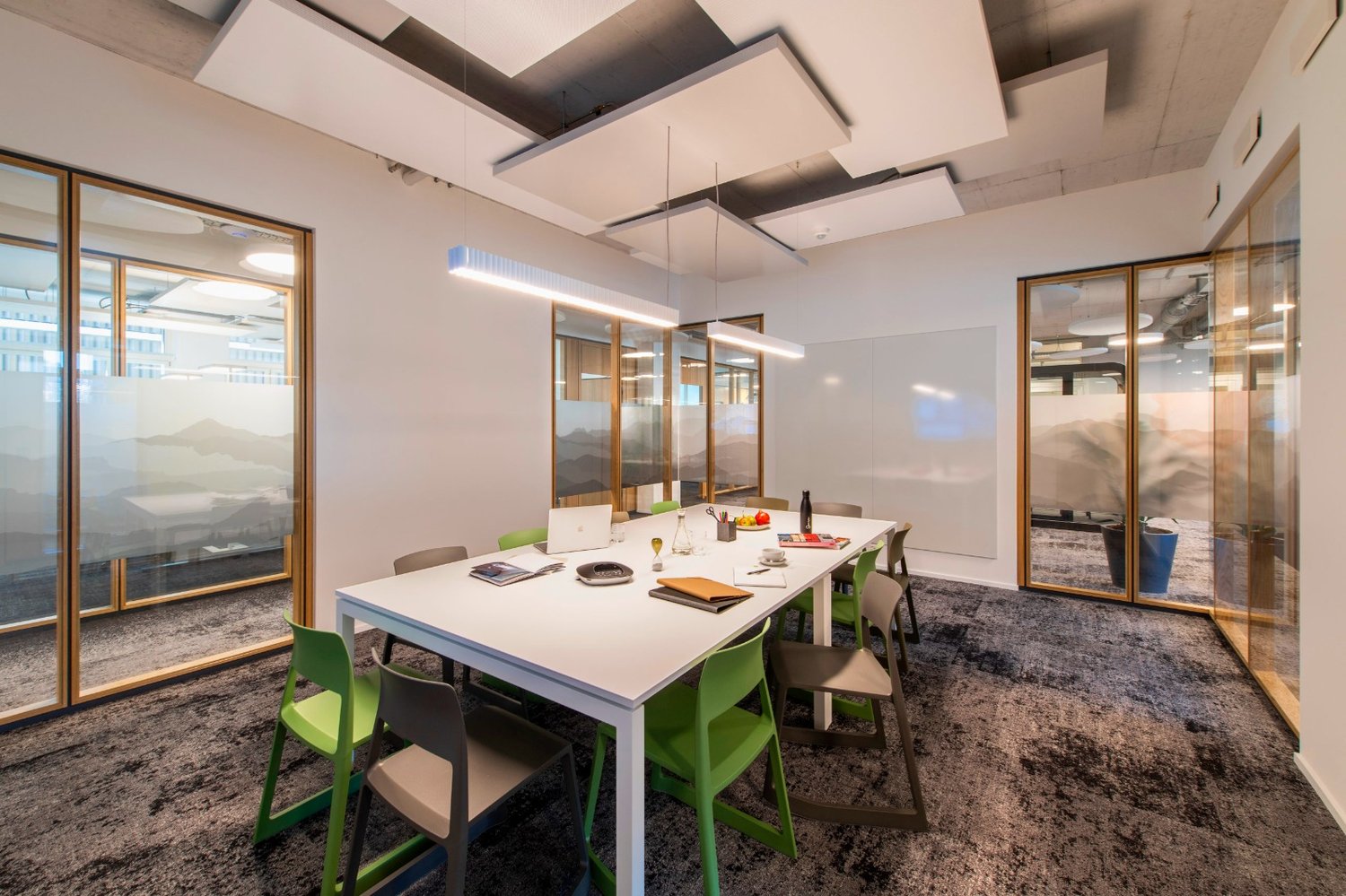 Large meeting room with white walls, modern lighting fixtures, a long white table, green and black chairs, and a glass wall overlooking the outdoors.