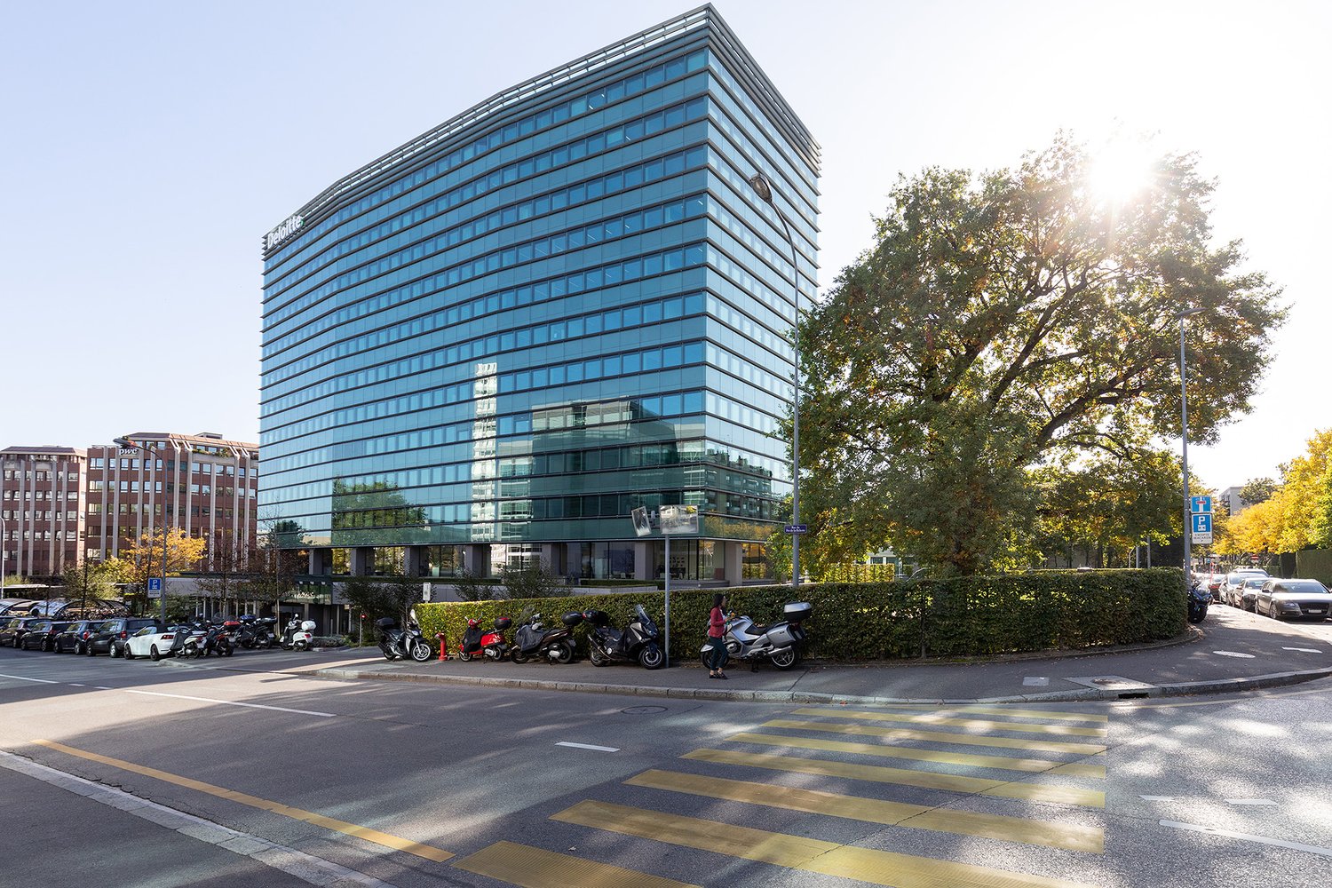 Large modern glass office building with multiple floors, surrounded by trees and parked vehicles on the street in front of the building