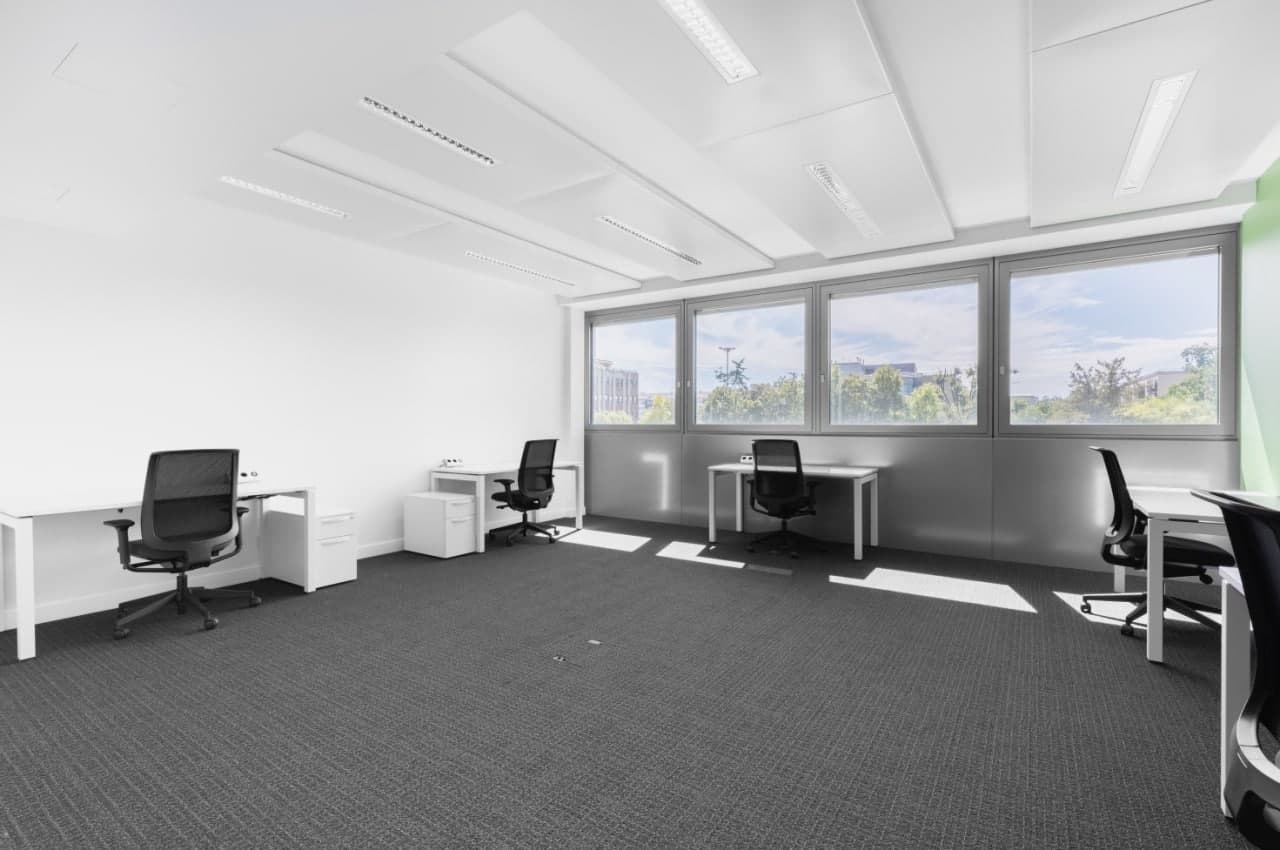 Large open office space with white walls, gray carpeted floor, multiple desks and office chairs, and large windows providing natural light and a view of the outdoors.