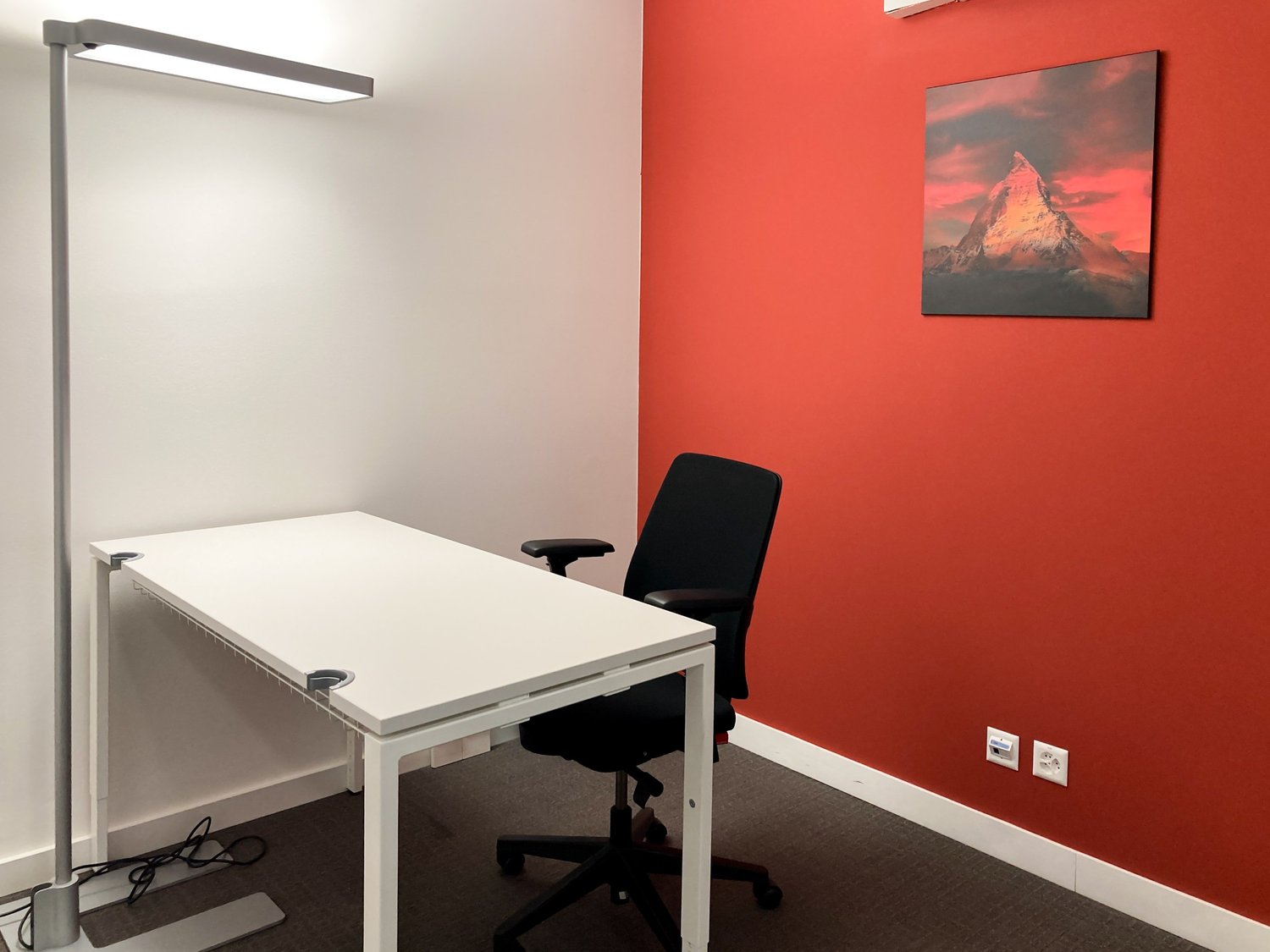 The image shows a small office space with a white desk, a black office chair, and a tall lamp. The walls are painted in a bright red color, and there is a framed artwork depicting a mountain landscape on the wall.