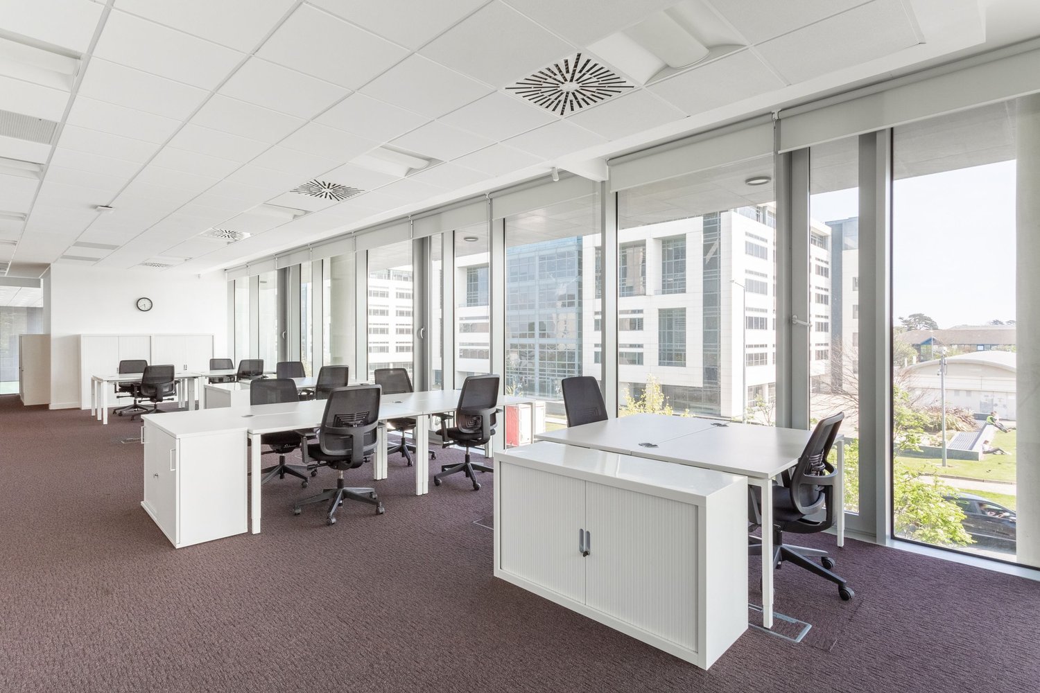 Large open office space with multiple workstations, desks, and chairs. The room has large windows providing a view of the surrounding buildings and landscape. The space appears to be well-lit with ceiling lights and has a modern, minimalist design.