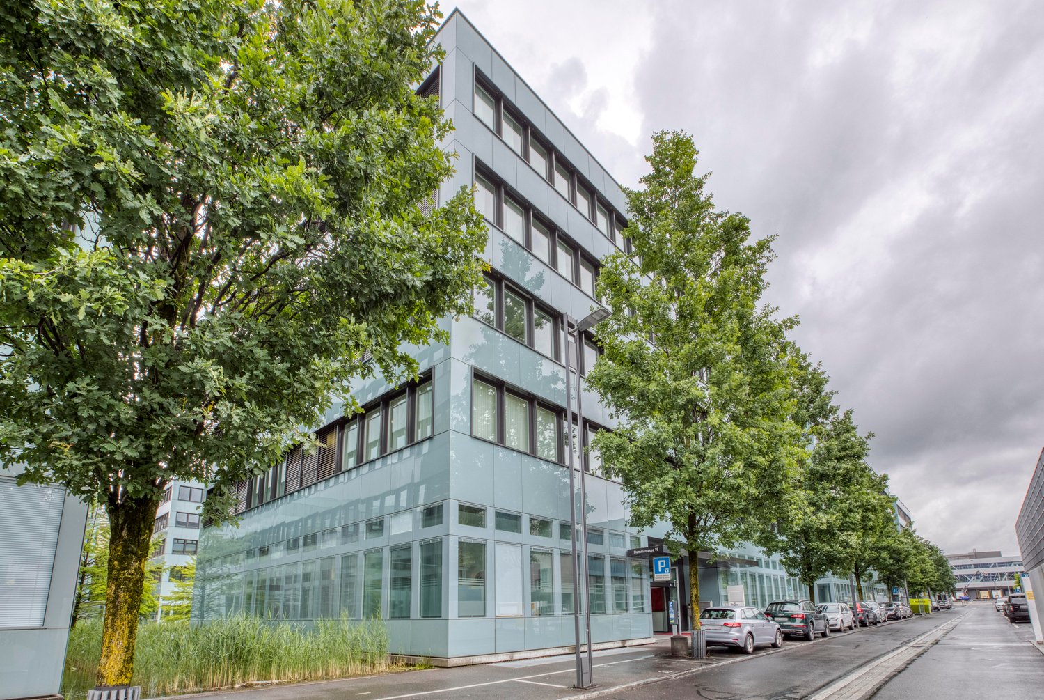 A modern, multi-story office building with a glass facade surrounded by trees and greenery. The building has several floors with large windows and a parking area in front.