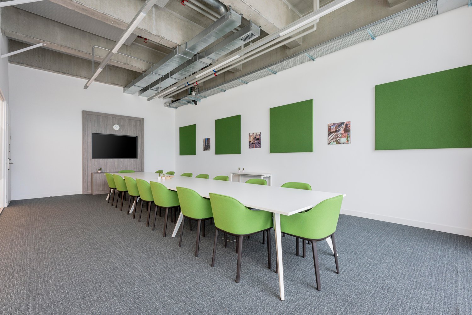 large table with chairs, carpeted floor, white walls, ceiling with metal ducts, monitor on wall, gray metal poles