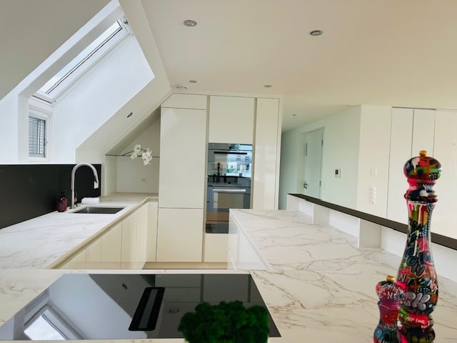 Modern kitchen with white marble countertop, white cabinets, black induction stove, stainless steel sink with faucet, a large open space, a skylight, and ceiling lamps.