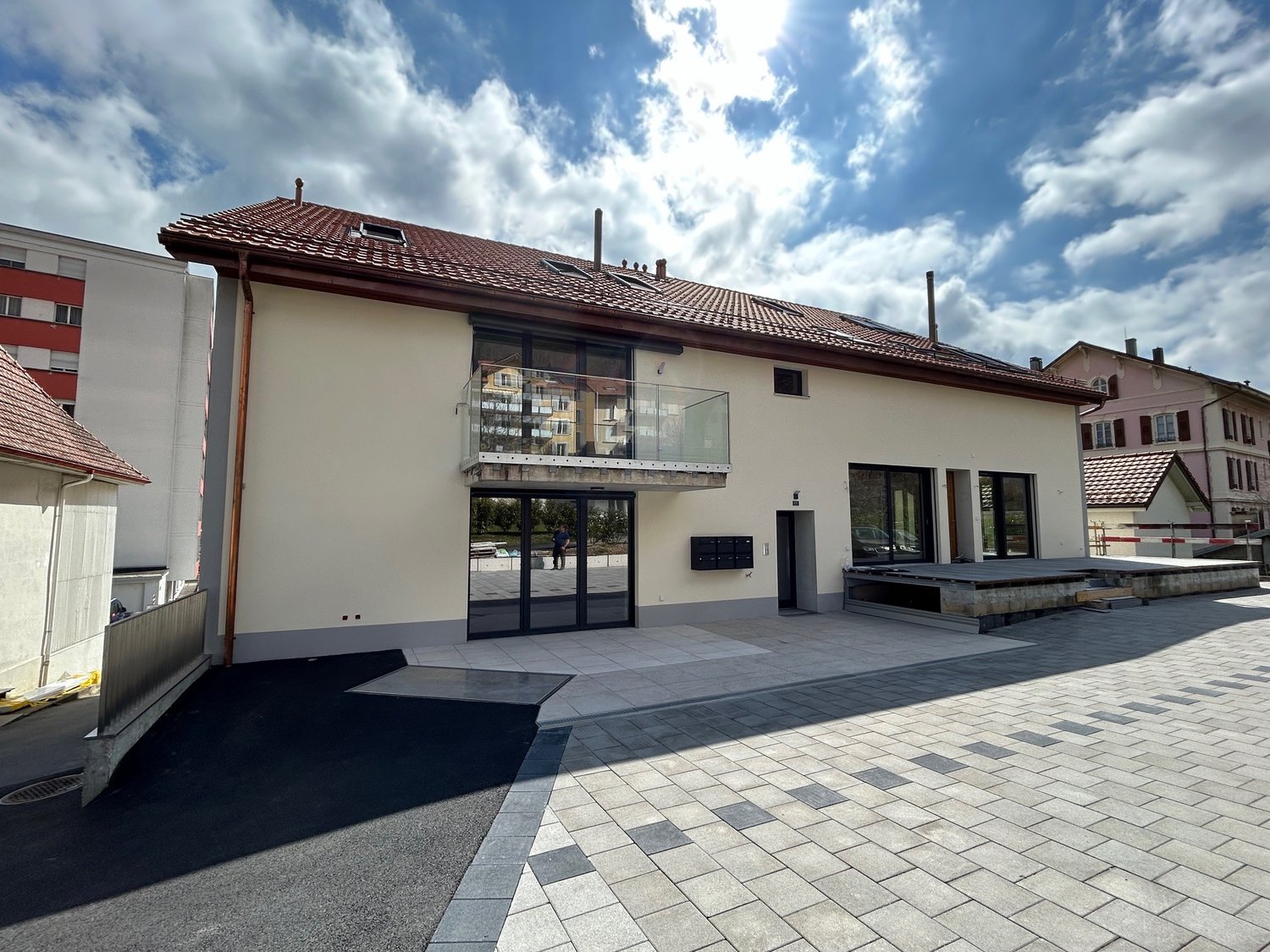 2-story building with a red tiled roof, large windows, and a balcony on the upper floor. The building has a modern, minimalist design with a paved outdoor area in front.