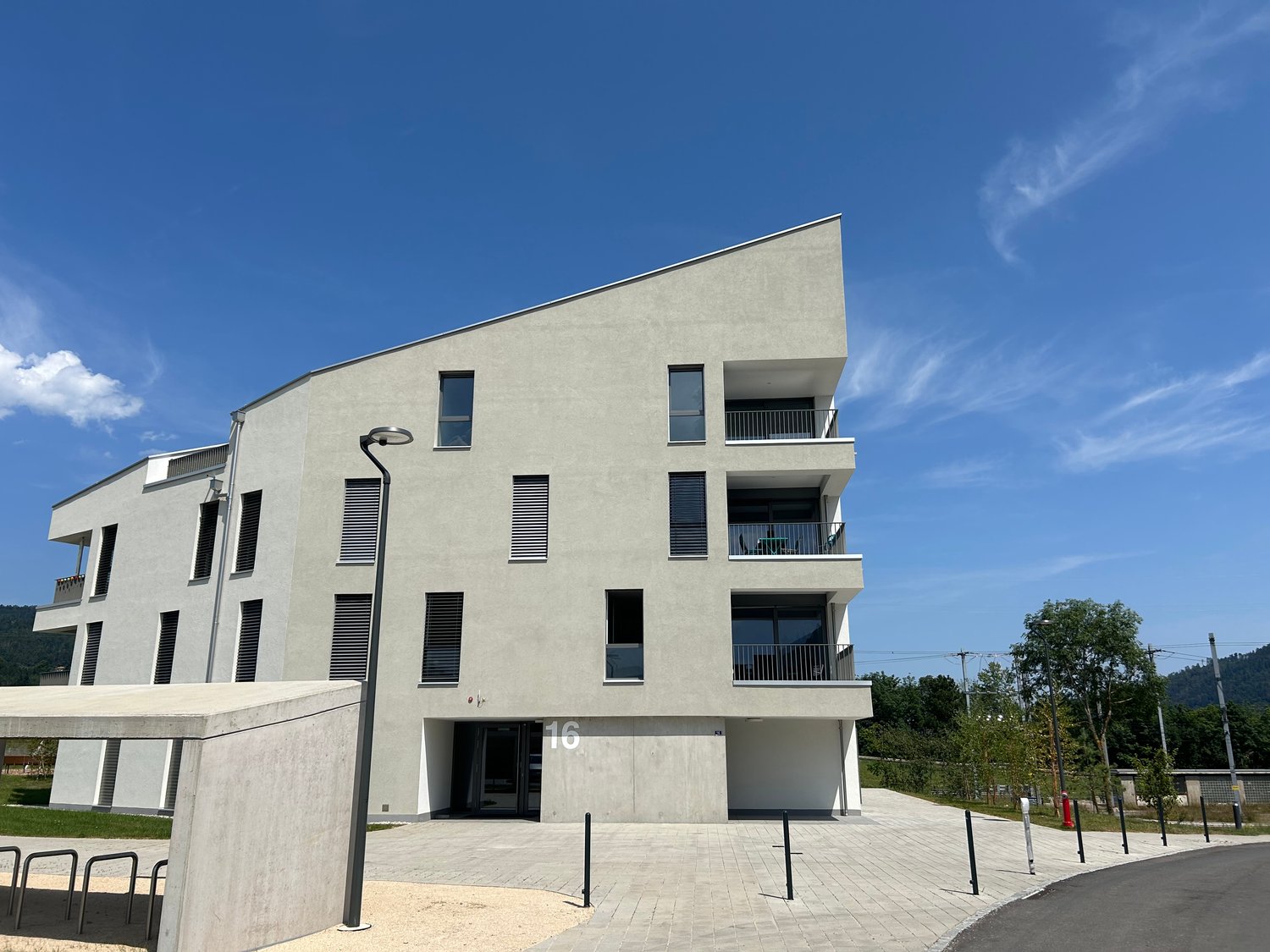 3-story modern apartment building with a gray exterior, large windows, and balconies on each floor. The building has a clean, minimalist design and is surrounded by a paved courtyard area.