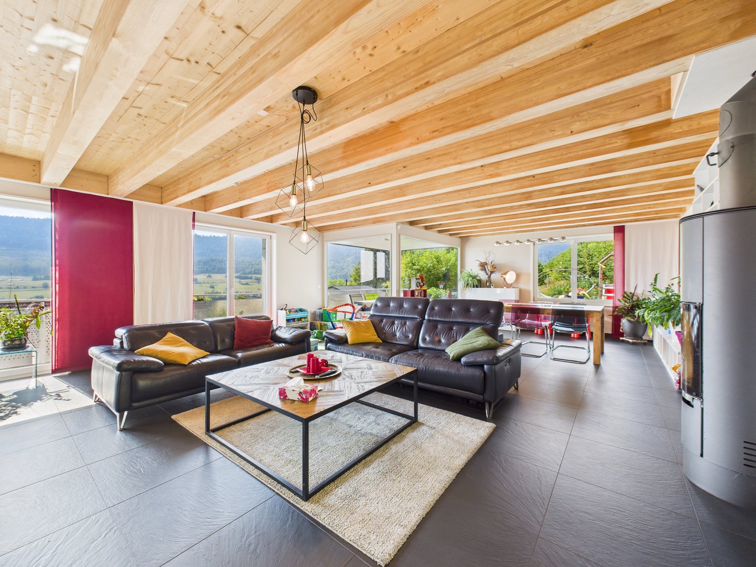 Open-plan living room with wood beams on the ceiling, red walls, two leather sofas, a center table, a fireplace on the wall, and large windows with an exterior view. A dining table and chairs can be seen in the adjacent dining area. There are plants on th