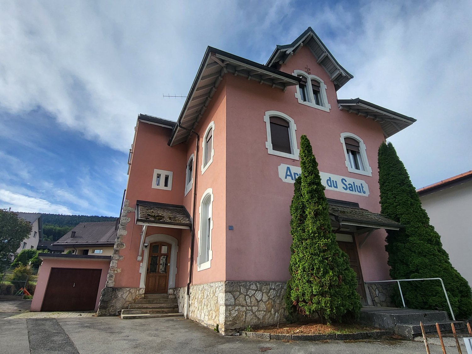 A two-story pink building with arched windows, a stone foundation, and a garage. The building has the name 'Arc du Salut' written on it and is surrounded by trees and shrubs.