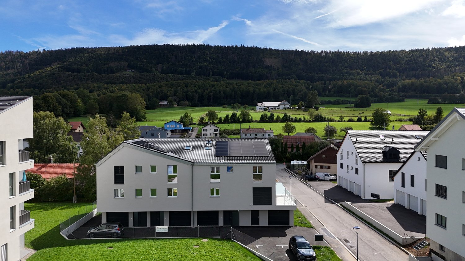 A gray building with a flat roof that has many windows, solar panels, a garage, cars parked in front of the house, a mountain and trees in the distance.