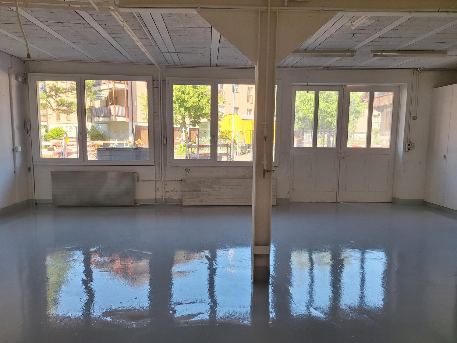 Empty room, high ceiling, multiple large windows, concrete floor, large central pillar, exterior view visible through windows