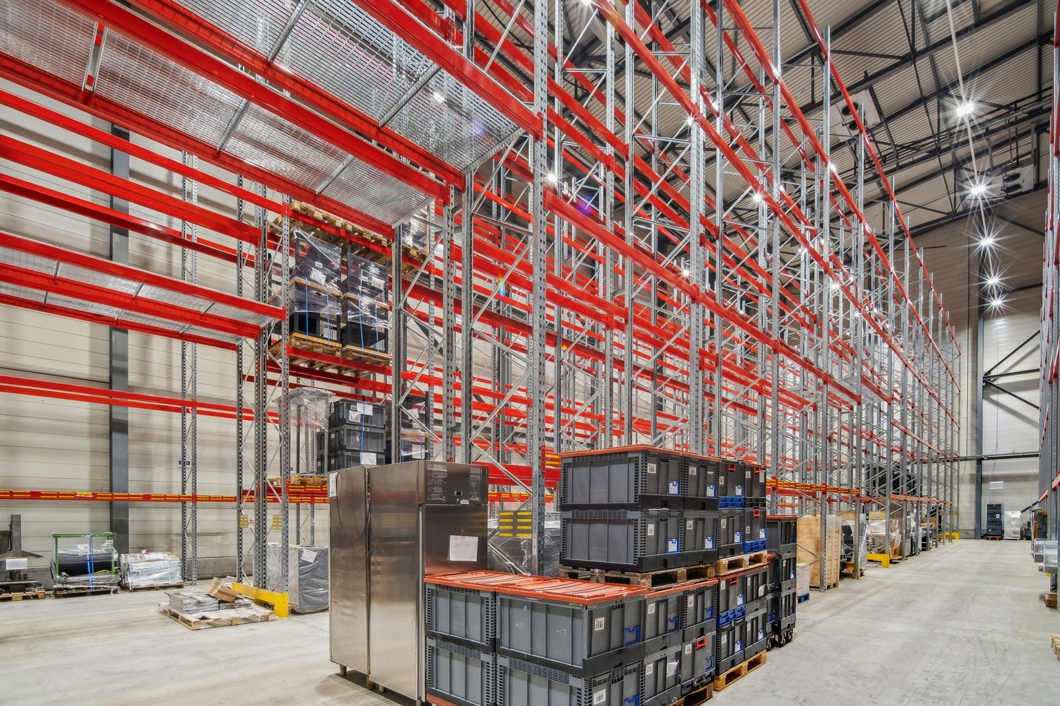 large warehouse with red and silver metal shelving, multiple stacked boxes, large silver refrigerator, concrete floor