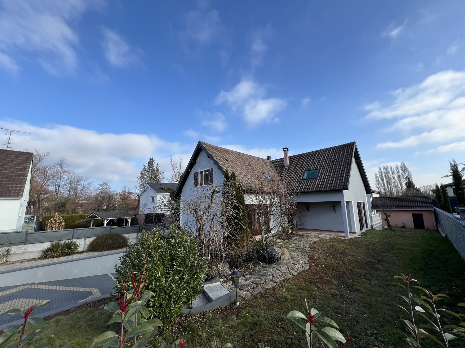 Two-story house with a tiled roof, surrounded by a well-maintained garden with various plants and shrubs. The house has large windows and a balcony or terrace visible.