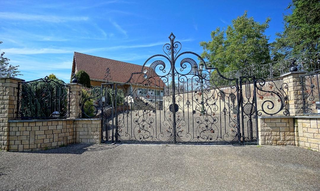 Two-story house with a red tile roof, surrounded by a decorative wrought-iron gate and stone walls. The property has a driveway leading to what appears to be a garage or carport.