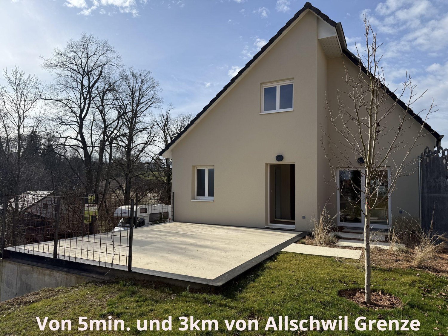 2-story house with beige exterior, black roof, large windows, concrete patio, fenced yard, and trees in the background