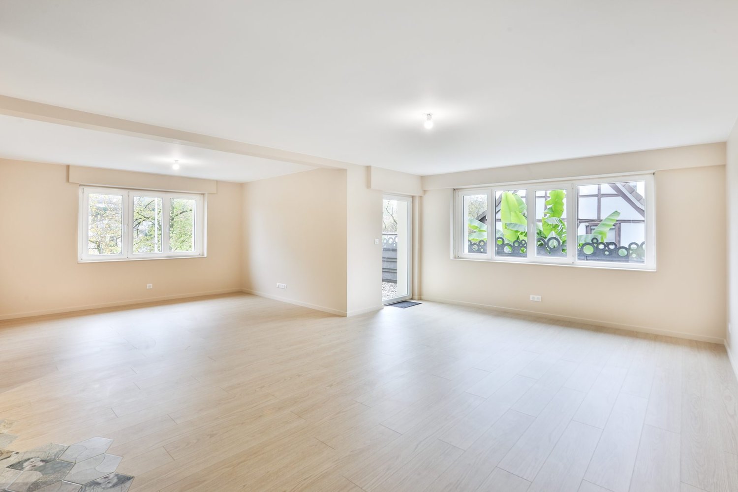 Spacious open-plan living area with large windows, hardwood flooring, and neutral wall colors. The room appears to be a combination of a living room and dining area, providing ample natural light and a bright, airy atmosphere.
