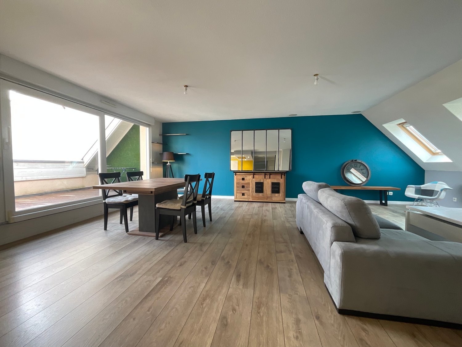 wooden floors, teal accent wall, dining table and chairs, living room sofa, round mirror, sideboard, table lamp, wooden bench, attic ceiling