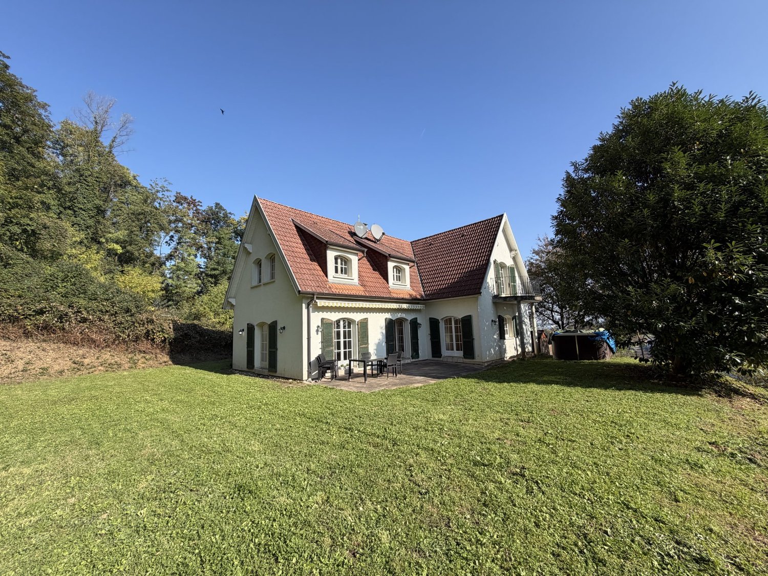 Detached white house, red roof, green shutters, garden table and chairs, large front yard