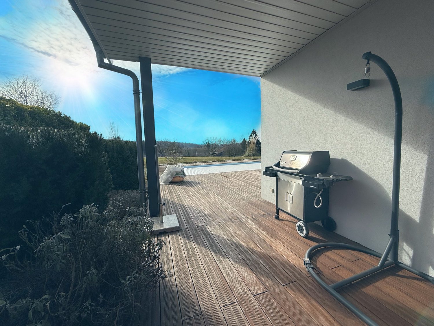 Covered outdoor patio with wooden deck, gas grill, and a view of the swimming pool and surrounding landscape.