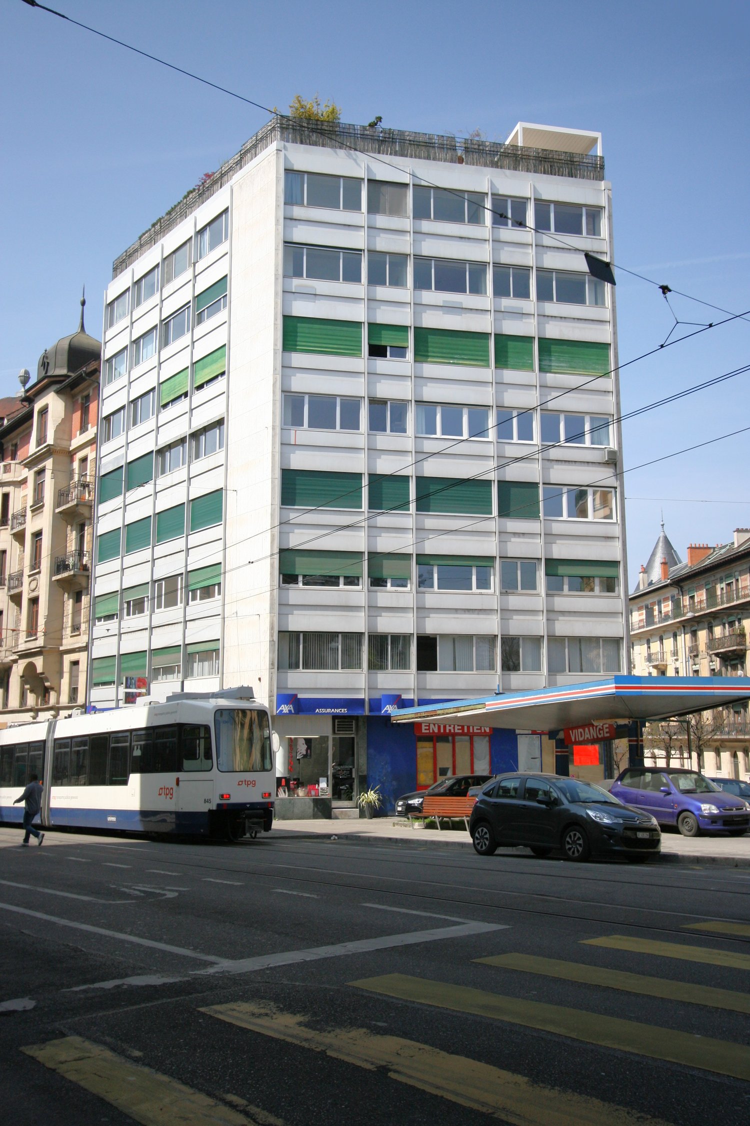 Multi-story commercial building with white exterior and green accents, balcony on the top floor, tram passing by on the street, other vehicles parked in front of the building