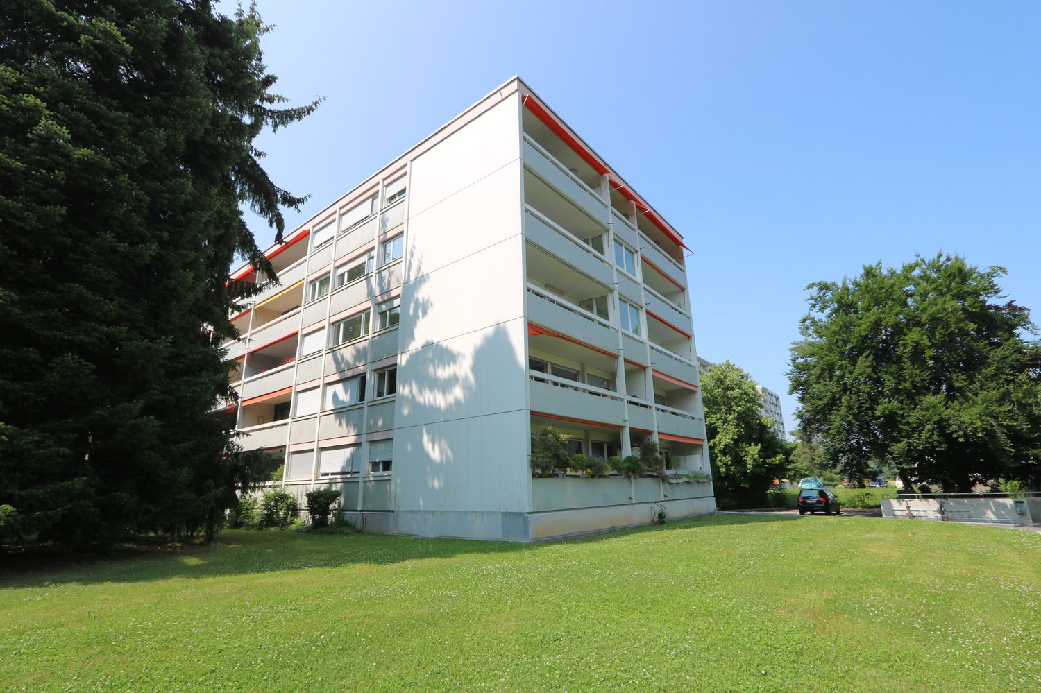 4-story apartment building with white exterior, red accents, and balconies. The building is surrounded by a grassy lawn and trees.