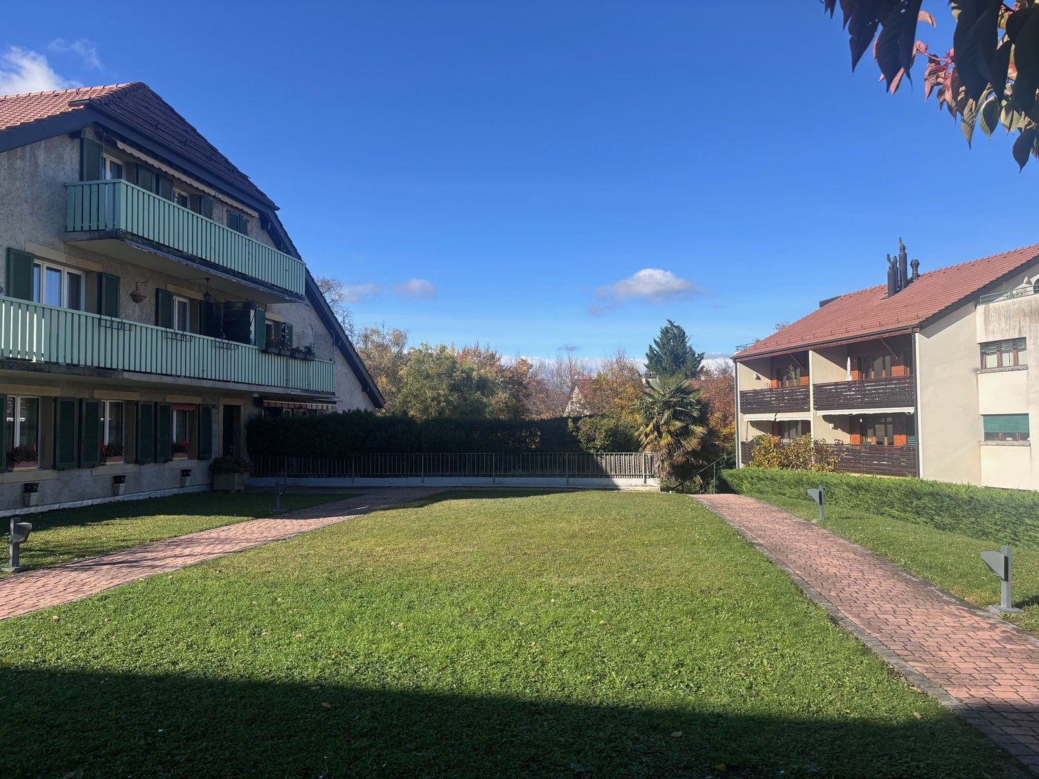 Two-story building with brown roofs, balconies, windows, and green shutters. A garden with a brick path in the front.