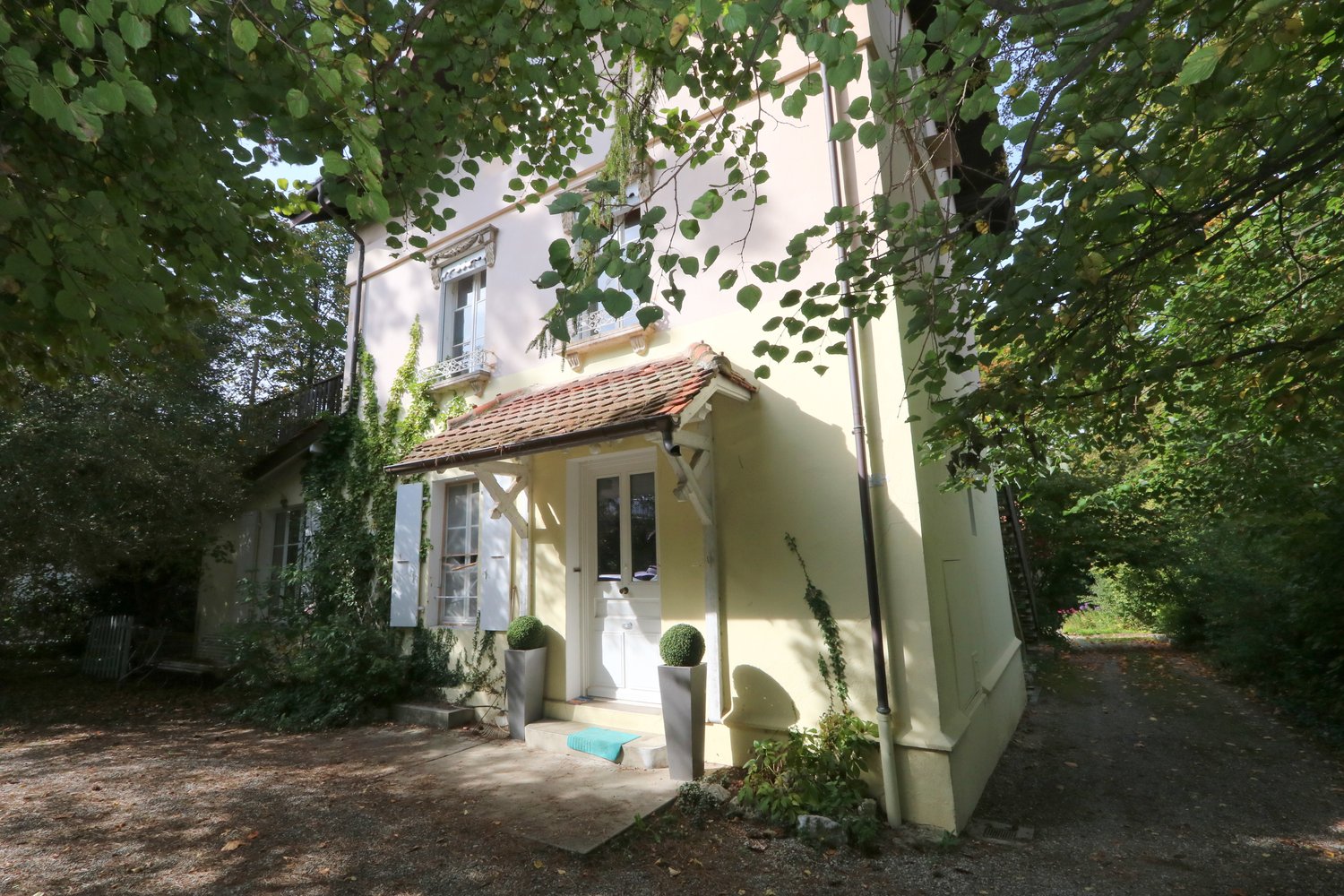 A two-story house with a yellow exterior and a brown roof. The ground floor is raised with concrete steps leading to the entrance. The windows are adorned with white shutters. The house is surrounded by a yard with trees and shrubs. There is a pathway lea