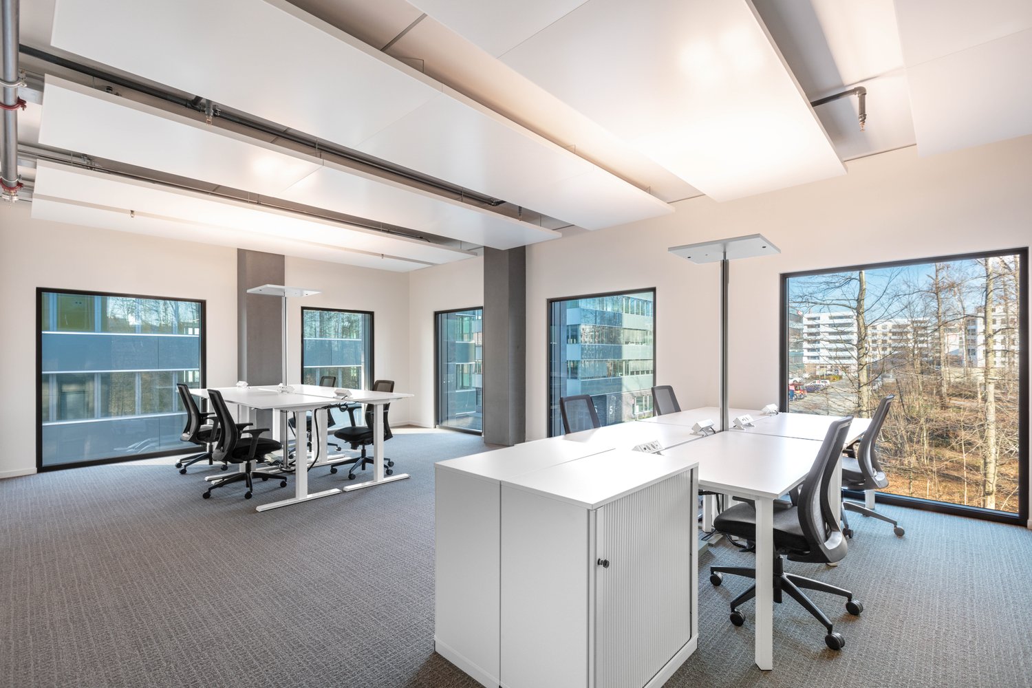 Spacious open-plan office with large windows providing a view of the surrounding trees and buildings. The room features white desks, black office chairs, and a central cabinet. The ceiling has exposed beams and lighting fixtures. The overall design appear