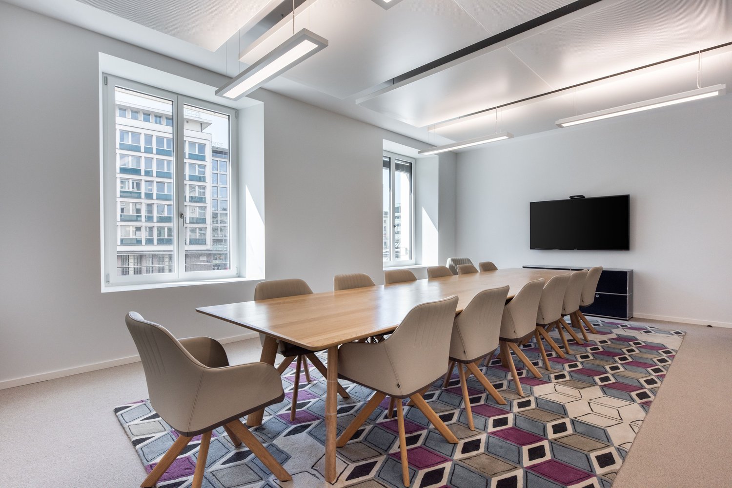 Large conference room with wooden table and chairs, modern lighting fixtures, large windows overlooking city buildings, and a patterned area rug on the floor.